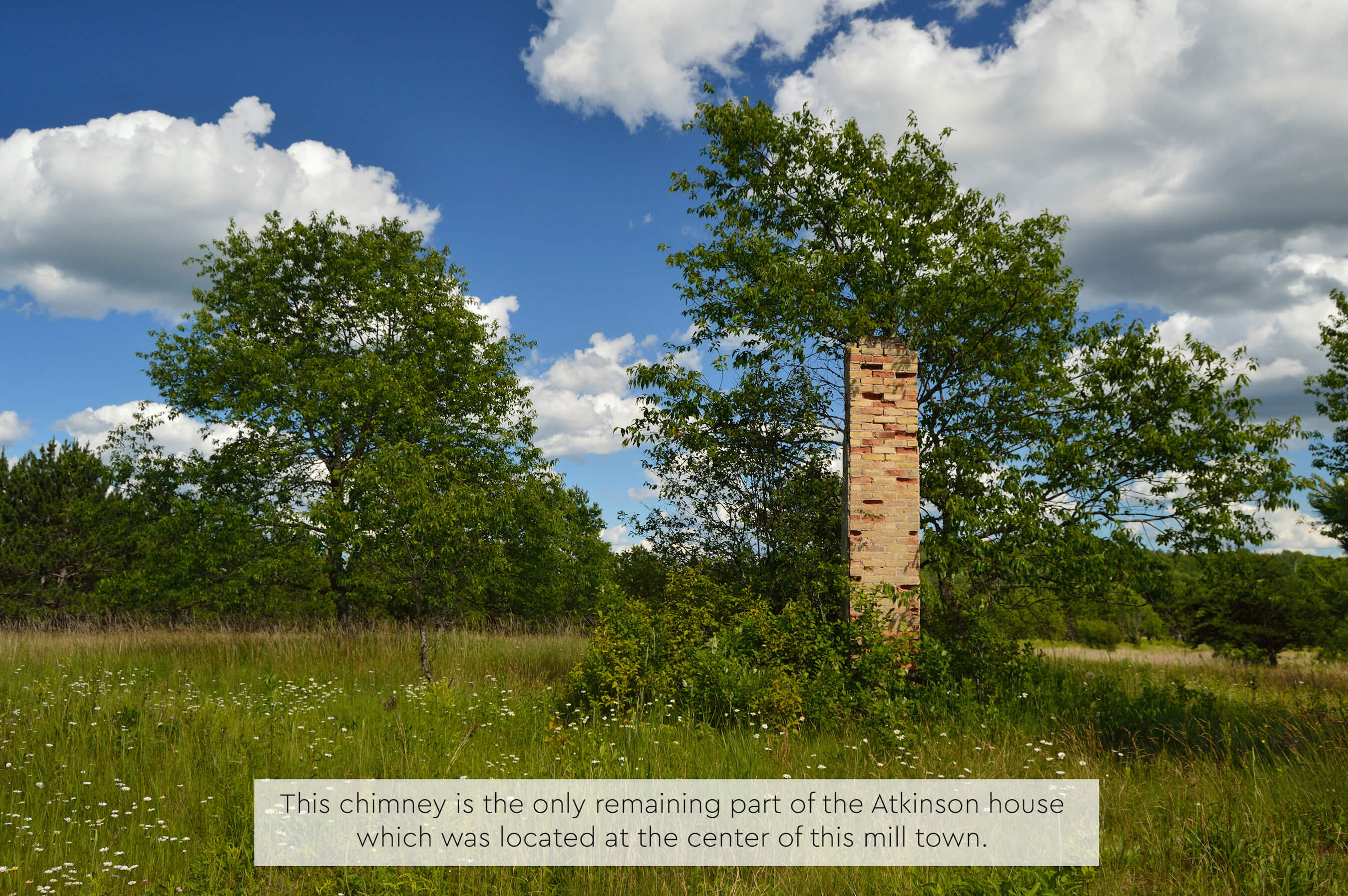An old brick chimney alone in a grassy field surrounded by green trees under a blue sky.