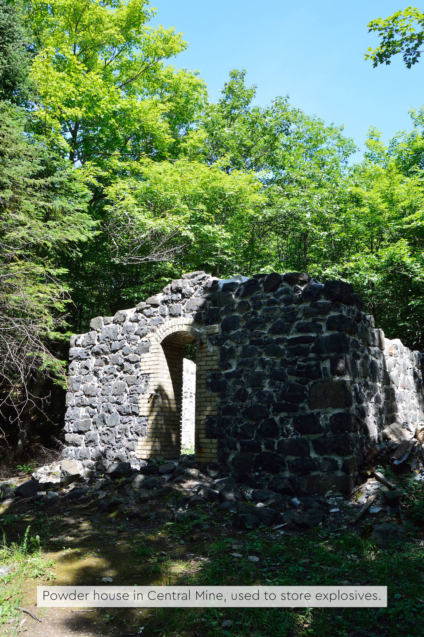 Stone ruins of a powder house with arched openings, surrounded by green trees and forest shadows.