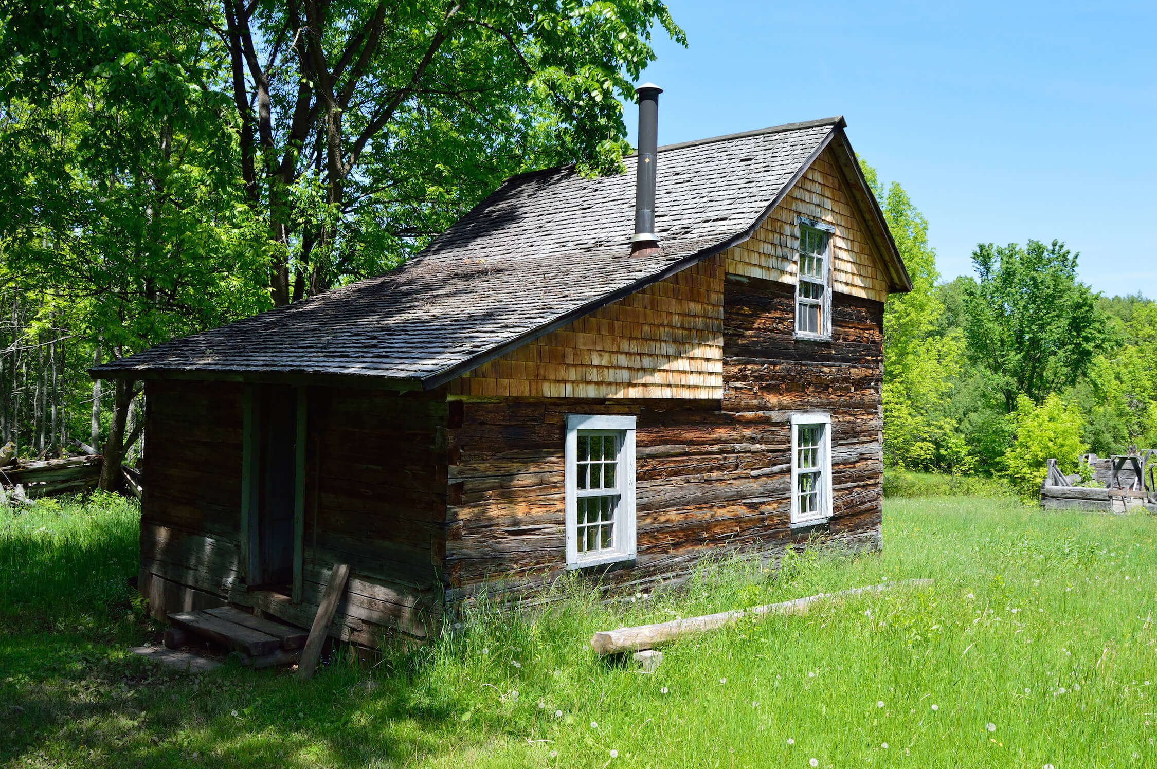 Historic log house with a steep roof and wooden shingles, surrounded by greenery.
