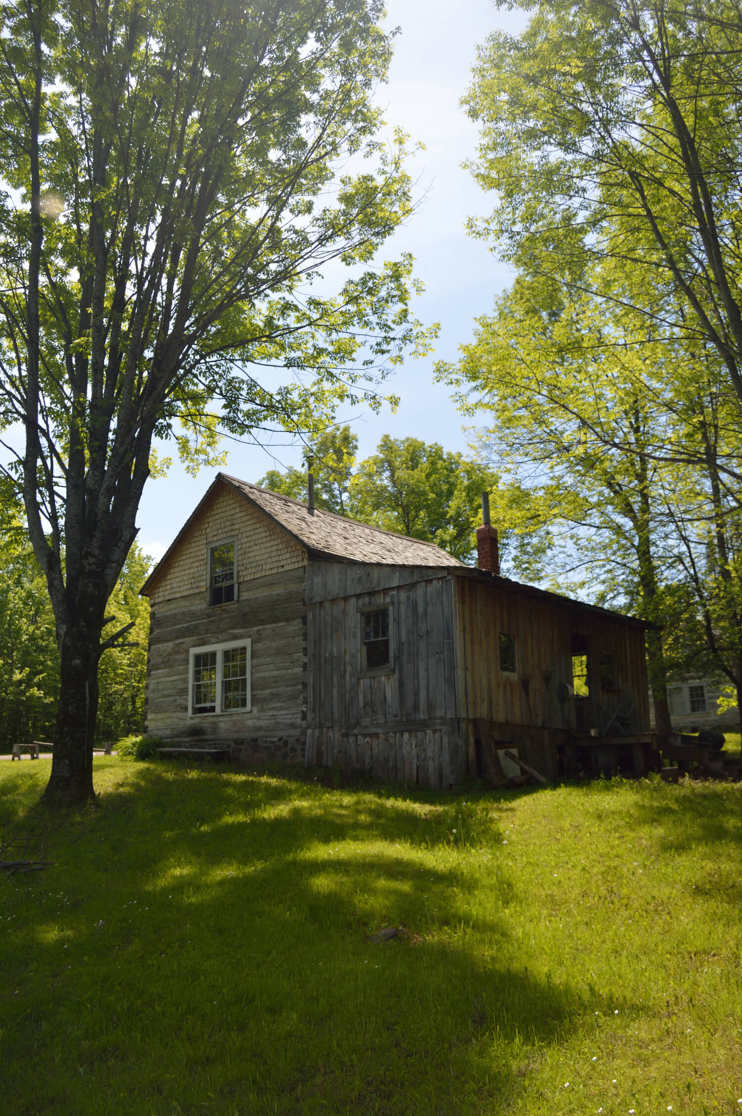 Old wooden house with a slanted roof surrounded by trees and grass on a sunny day.
