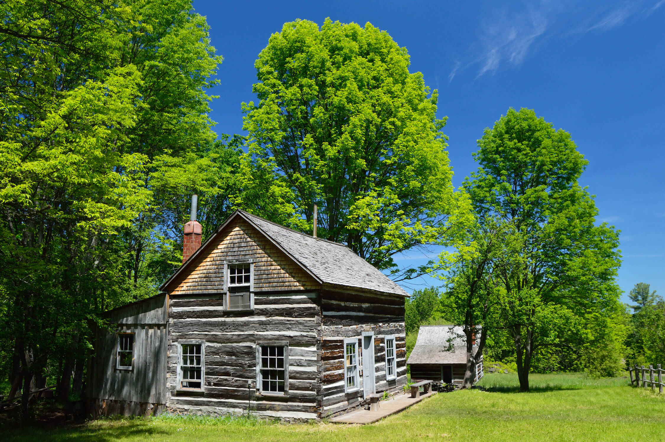 Restored log house with white-trimmed windows, surrounded by trees and greenery.