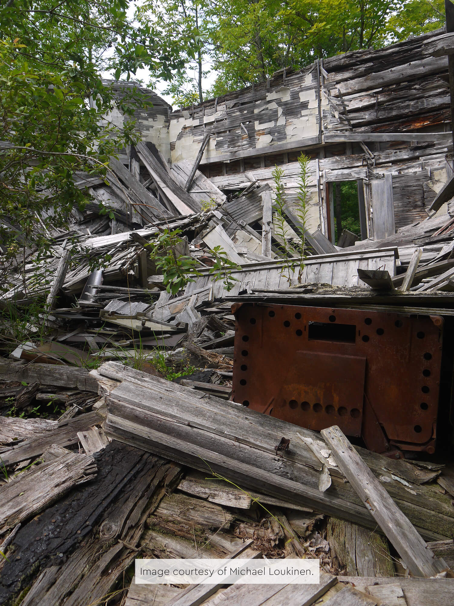Ruins of a collapsed wooden building with rusted metal stove in foreground.