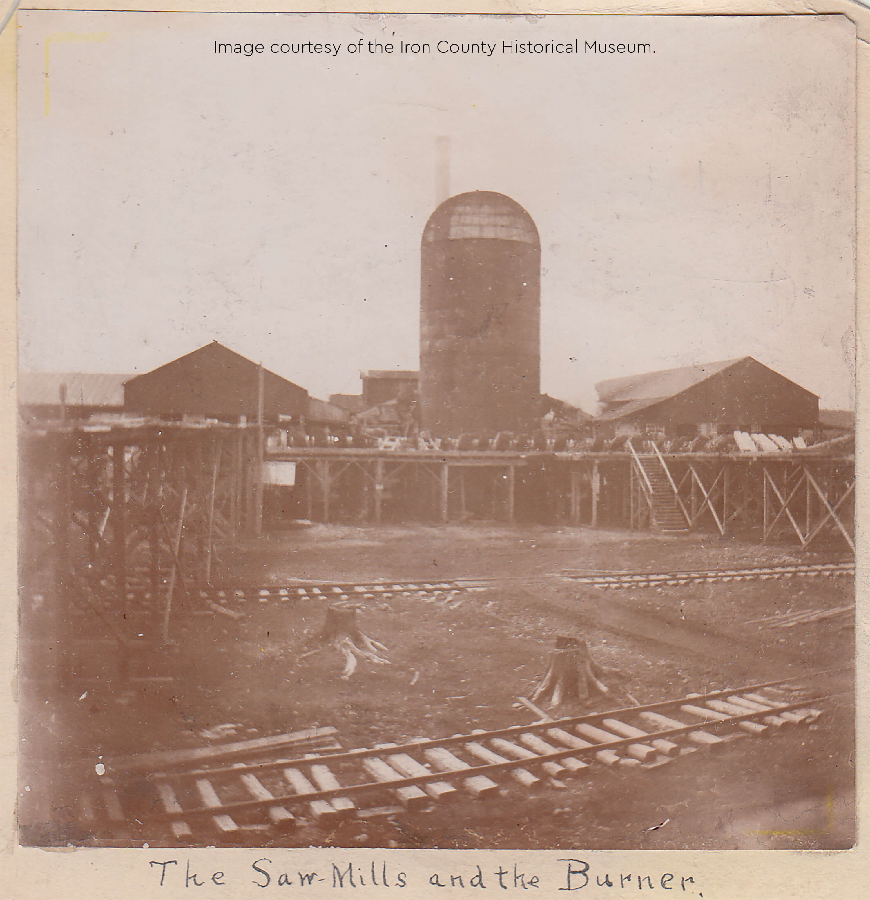 A historical photograph showing sawmills and a large cylindrical burner with wooden tracks.