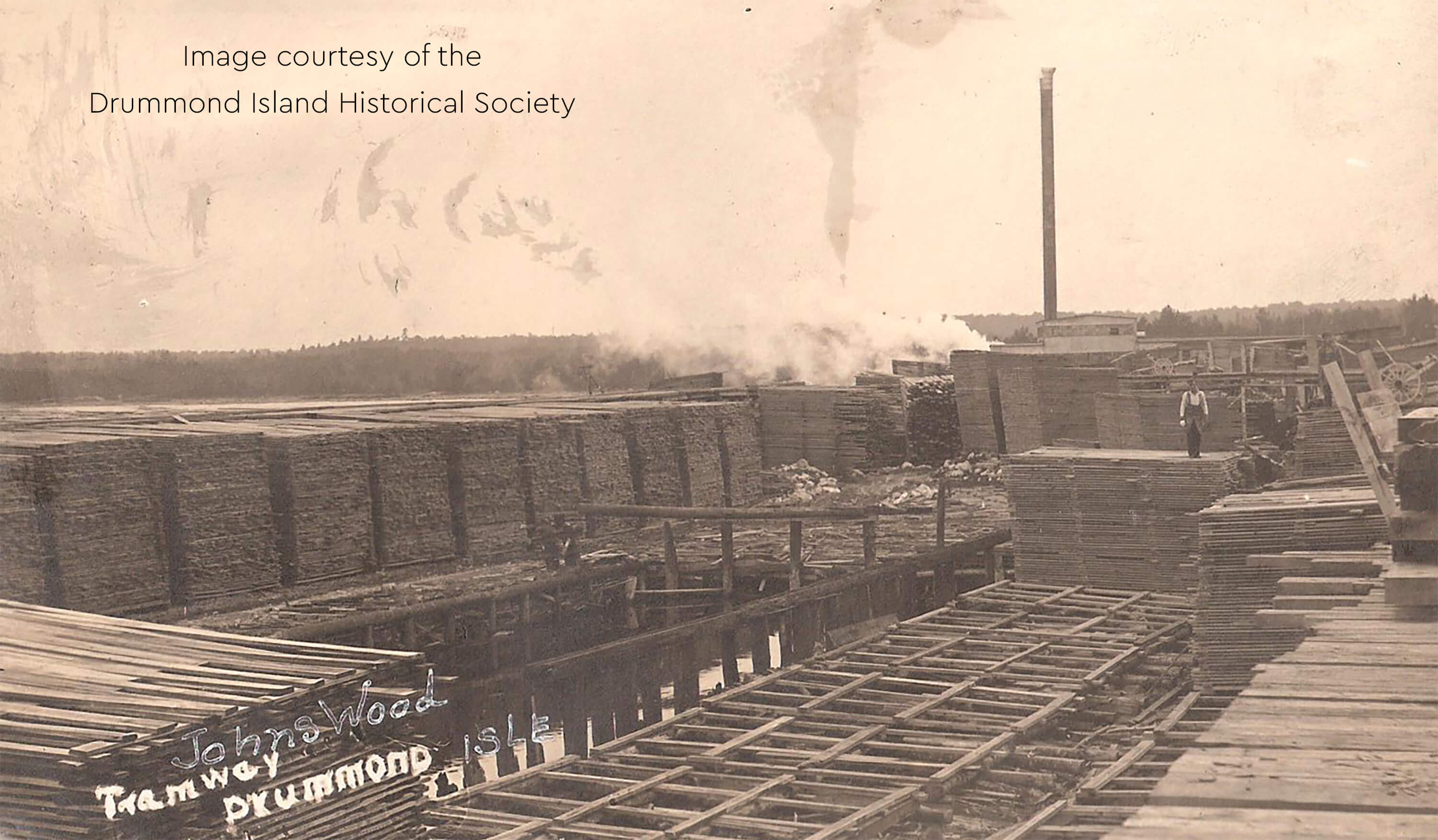 Historic image of stacked lumber at Johnswood with a tramway and smokestack.
