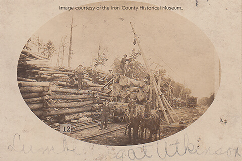 Workers stand among large stacks of logs with horses and machinery at a lumber yard in Atkinson.