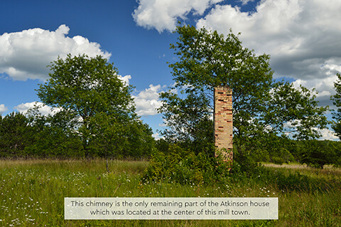 An old brick chimney alone in a grassy field surrounded by green trees under a blue sky.