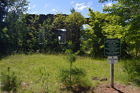 Stone ruins of Engine House No. 2 surrounded by greenery, with a warning sign in the foreground.