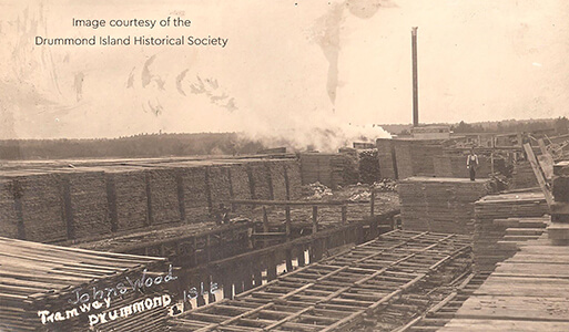 Historic image of stacked lumber at Johnswood with a tramway and smokestack.