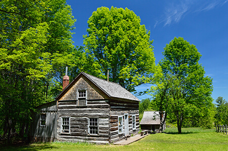 Restored log house with white-trimmed windows, surrounded by trees and greenery.