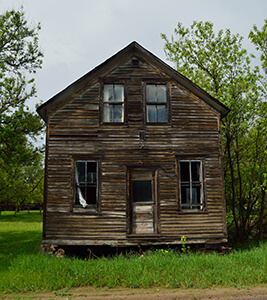 Old wooden house with broken windows and missing siding in a grassy area.
